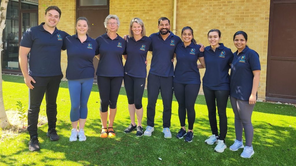 A group of people standing in a semi circle. They are all wearing blue MD Support Centre t-shirts. They comprise a mix of genders and ethnicities.