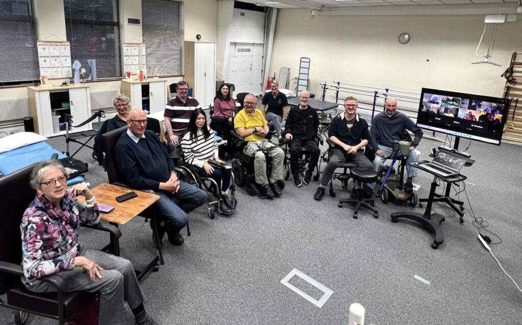 A group of people in chairs and wheelchairs sitting in a semi circle. There is also a TV with people who have joined online.
