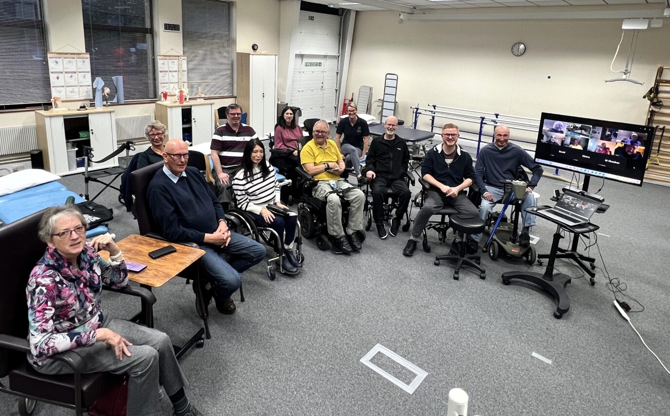 A group of people in chairs and wheelchairs sitting in a semi circle. There is also a TV with people who have joined online.