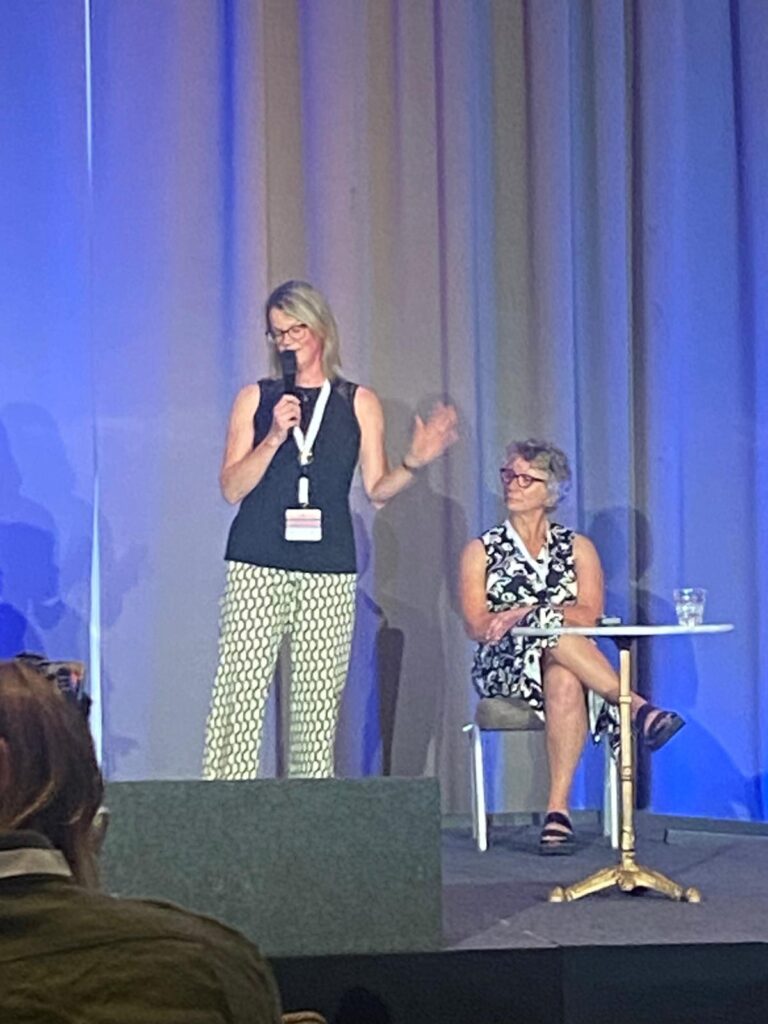 Lynn and Ulrike are on stage during their talk at FSHD Connect. Lynn is standing and speaking into a microphone. Ulrike is sitting down next to her.
