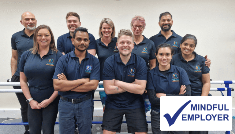 Members of MD Support Centre's therapy team standing in our centre. The Mindful Employer logo, featuring the words Mindful Employer next to a blue tick, has been superimposed over the image.
