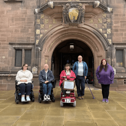 A group of people some in wheelchairs, others using scooters, and some standing, in front of a building in London