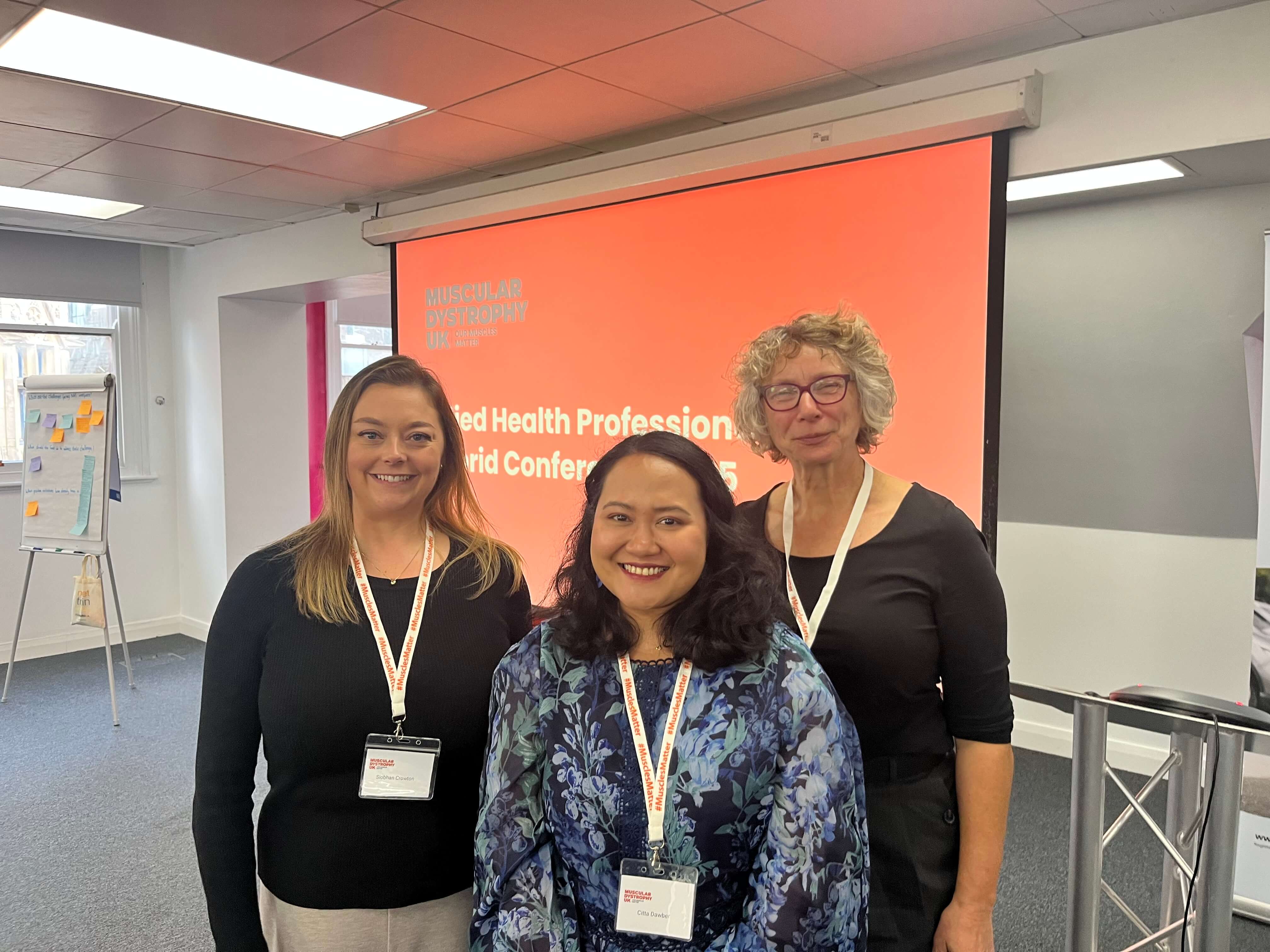 Physiotherapists Siobhan Crowton and Ulrike Uta with Citta Dawber from MDUK at the Allied Health Professionals' Conference. They are standing in front of an screen with MDUK's logo on it. All are smiling at the camera.