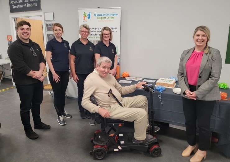 Daventry-launch-website Group photo at the Muscular Dystrophy Support Centre's Daventry clinic showing staff members standing alongside a wheelchair user, with a display table and centre banner in the background.