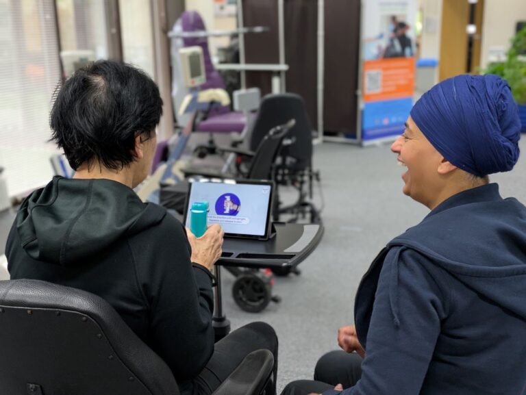 Two people sit in a rehabilitation space, engaged in hand therapy. They are looking at a tablet on a tray table displaying a graphic, with exercise equipment in the background