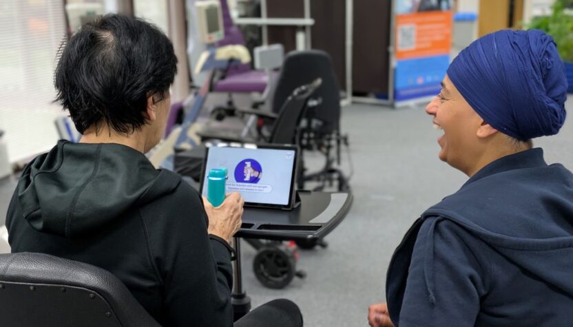 Two people sit in a rehabilitation space, engaged in hand therapy. They are looking at a tablet on a tray table displaying a graphic, with exercise equipment in the background