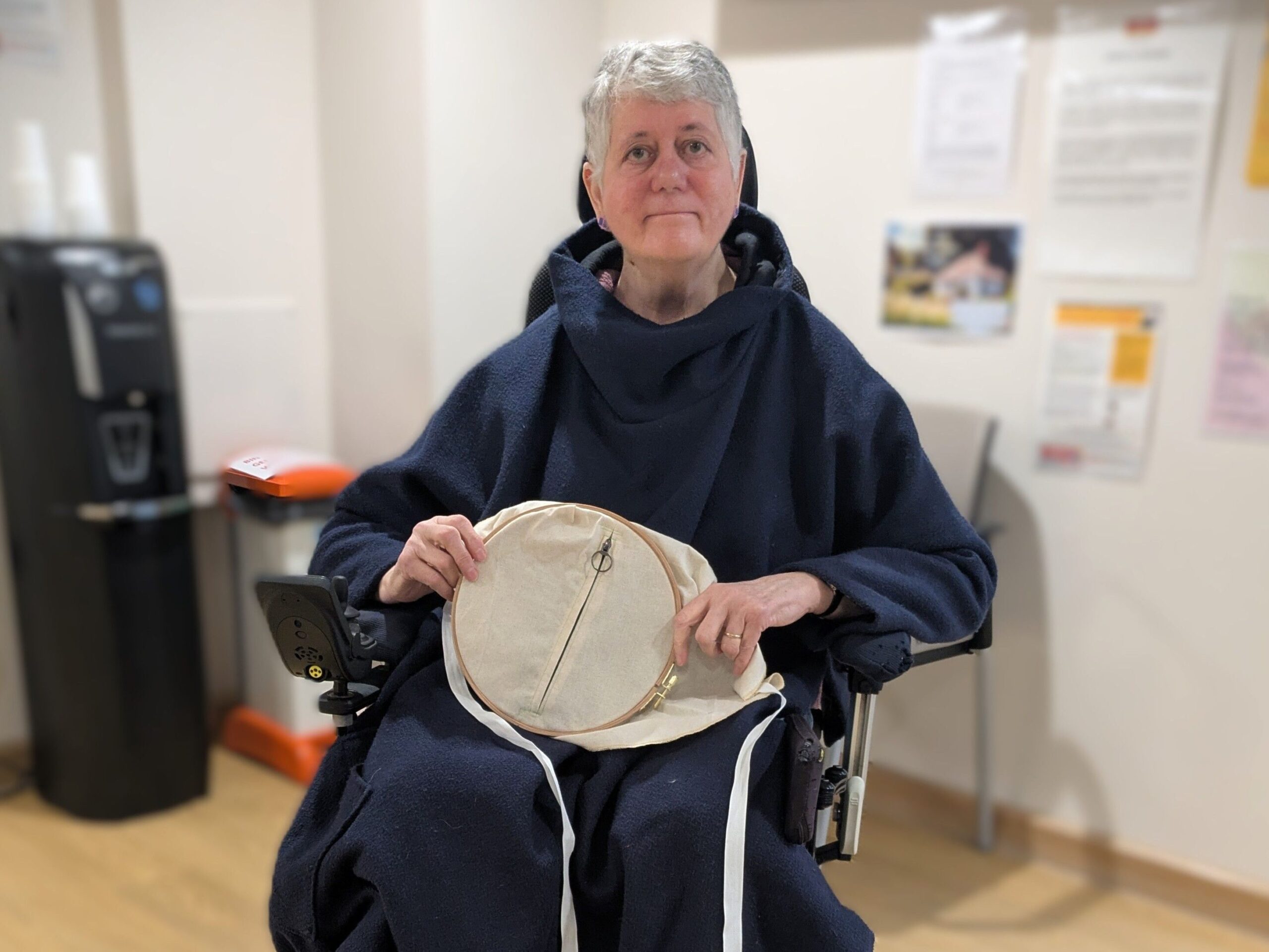 A person seated in a wheelchair holds a circular fabric frame with long fabric strips attached, in a room with notices on the wall and a water dispenser in the background.