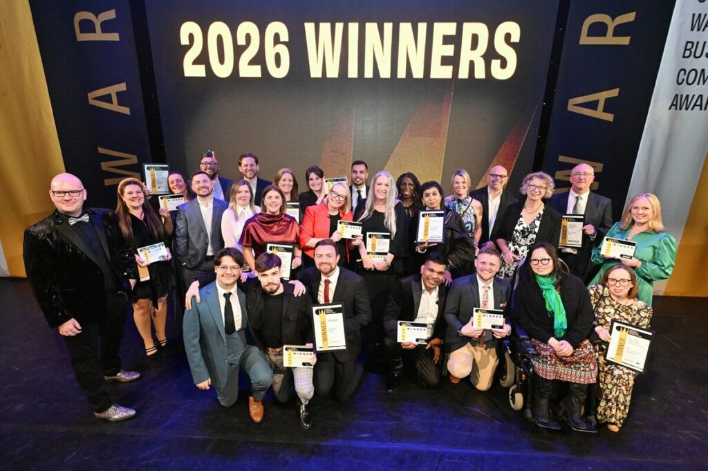 Large group of award winners posing on a stage beneath a screen reading “2026 Winners,” each holding an award certificate, with Coventry & Warwickshire Business and Community Awards branding in the background.