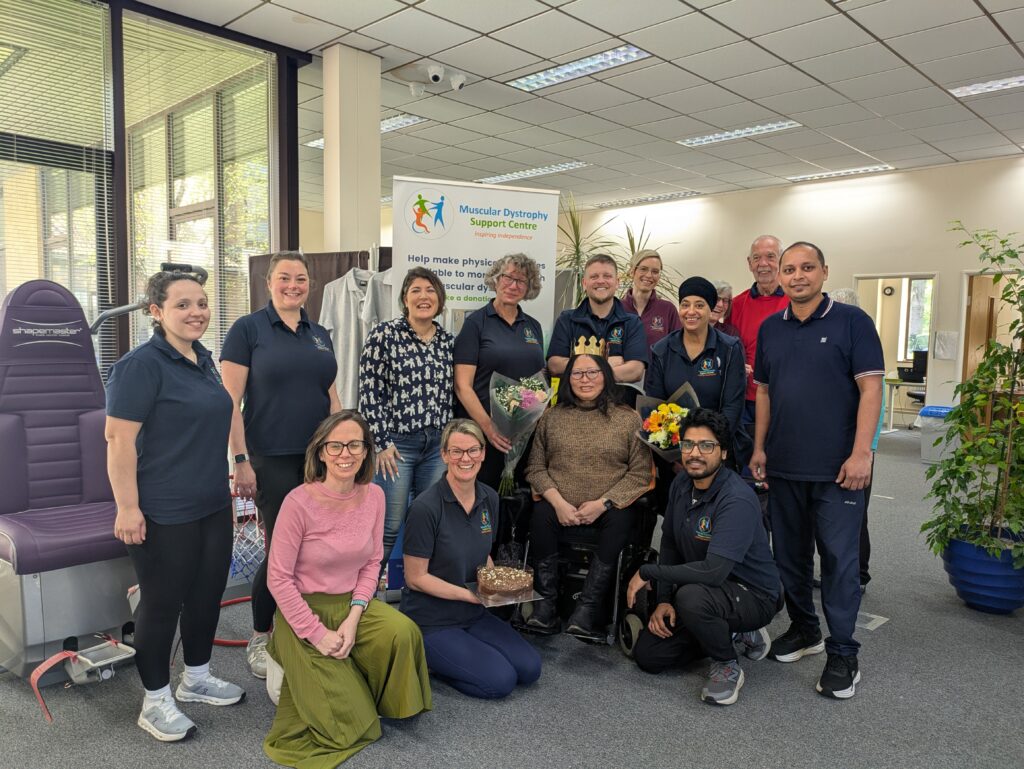 A group of people standing, sitting, and kneeling in MD Support Centre's office. In the middle, a woman is holding an award and other people are holding a cake and flowers
