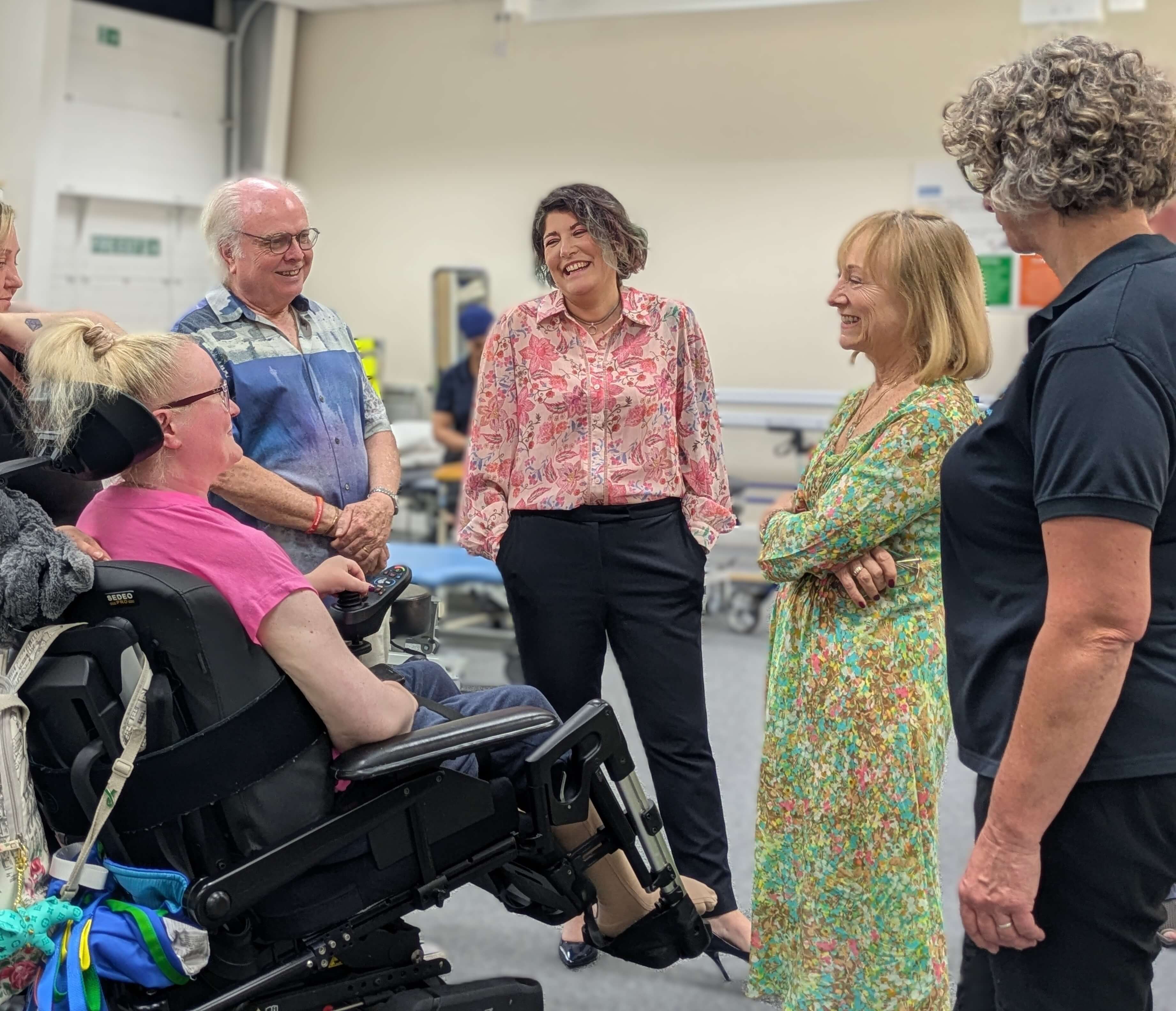 A group of adults stand in a therapy room speaking with a person using a powered wheelchair. The wheelchair user holds a control device, while others stand around in a semicircle. Therapy equipment and treatment tables are visible in the background.
