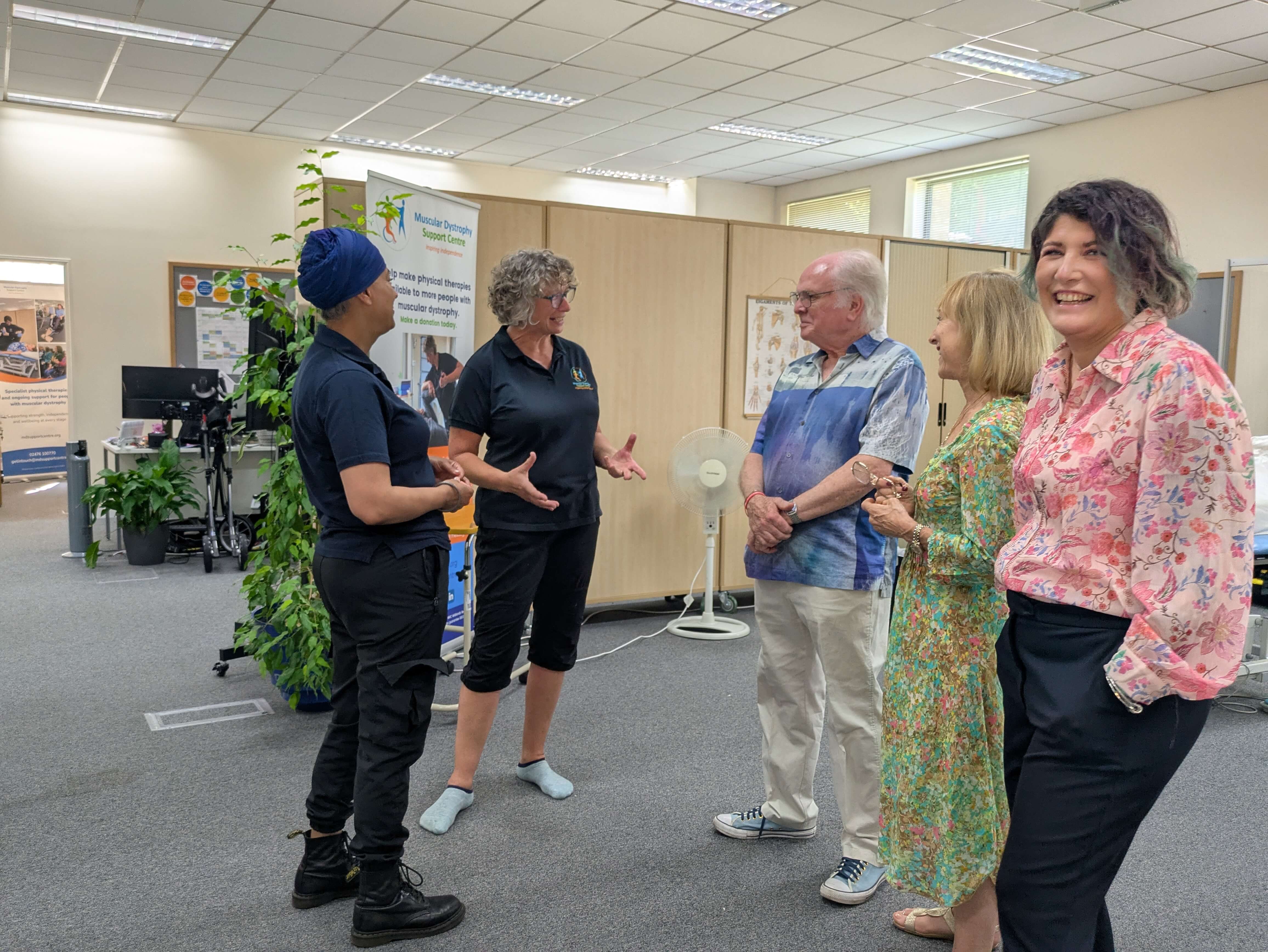 A group of five adults stand in a therapy room, talking together. Two staff members in dark polo shirts face three visitors. One person in a pink floral blouse looks toward the camera. The room includes posters, a banner for a muscular dystrophy support centre, desks, plants, and therapy equipment.