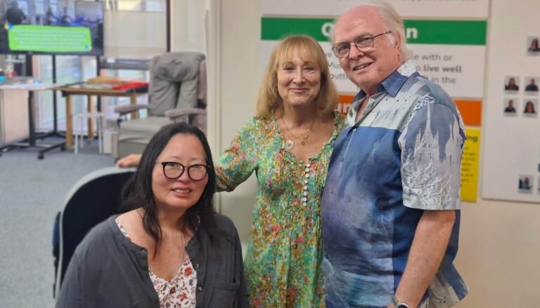 Three adults pose together in a clinical reception or therapy area. One person is seated wearing glasses, while two others stand beside them. Behind them are posters, a display board with text, and office furniture.