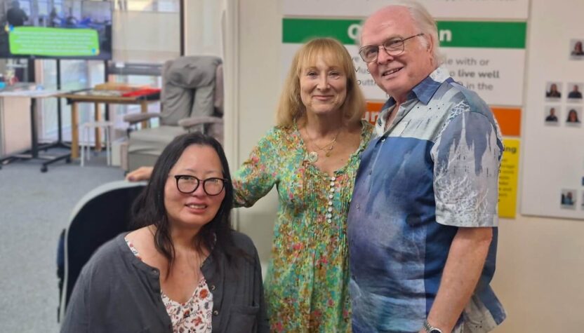 Three adults pose together in a clinical reception or therapy area. One person is seated wearing glasses, while two others stand beside them. Behind them are posters, a display board with text, and office furniture.