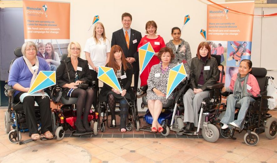 NMC Midlands is founded Group of people at a Muscular Dystrophy event, several using wheelchairs, holding colourful signs with charity banners in the background.