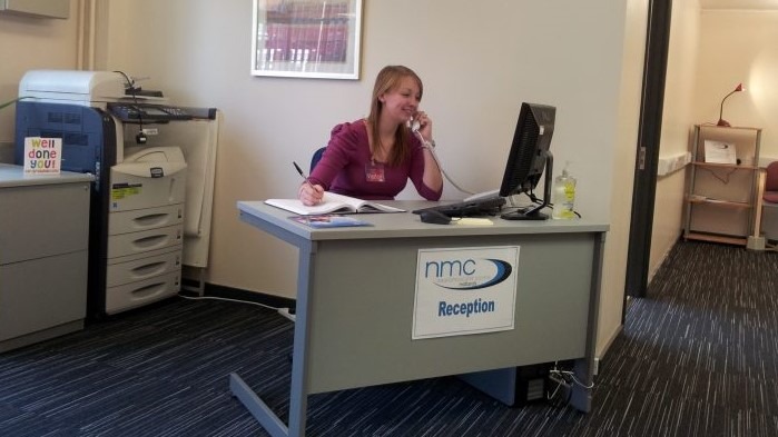 NMC Midlands opens its doors A woman behind a computer desk, talking on the telephone and writing in a notepad.