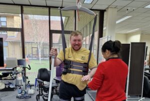 A therapist supports someone to walk using a ceiling tracked hoist and walking stick.