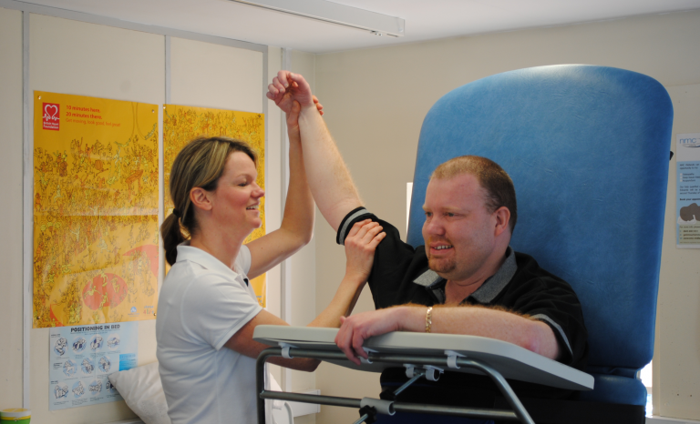 lynn-full-768x466 Physiotherapist supporting a person using a standing aid during a therapy session.