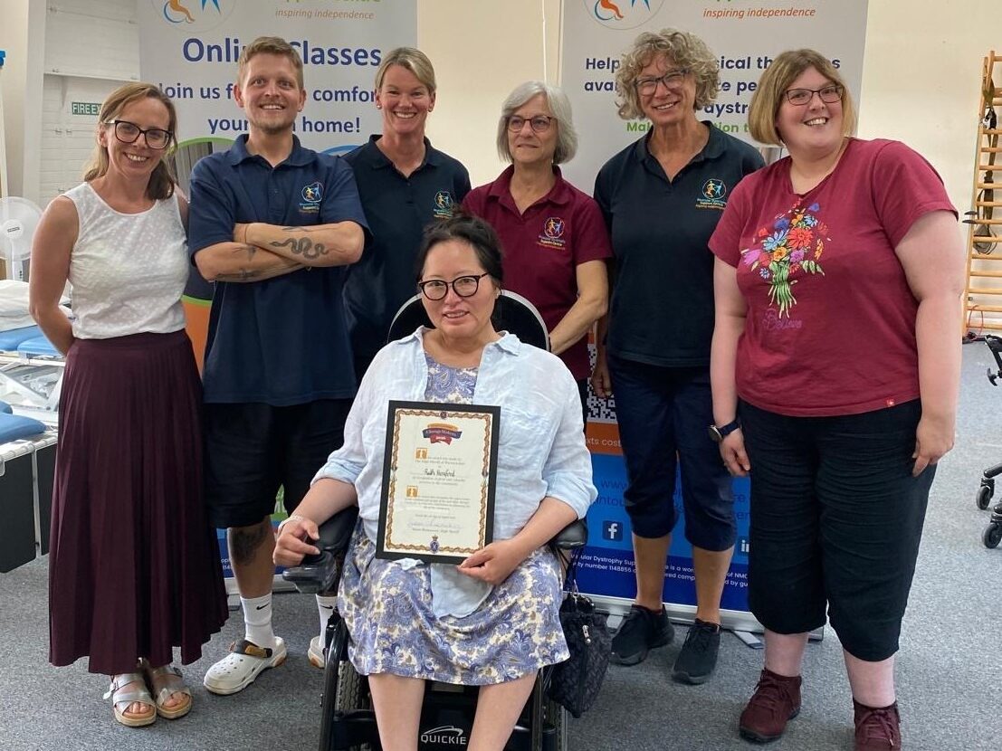 A group of people standing and one person sitting in a powerchair in MD Support Centre's therapy area. One holds a certificate.